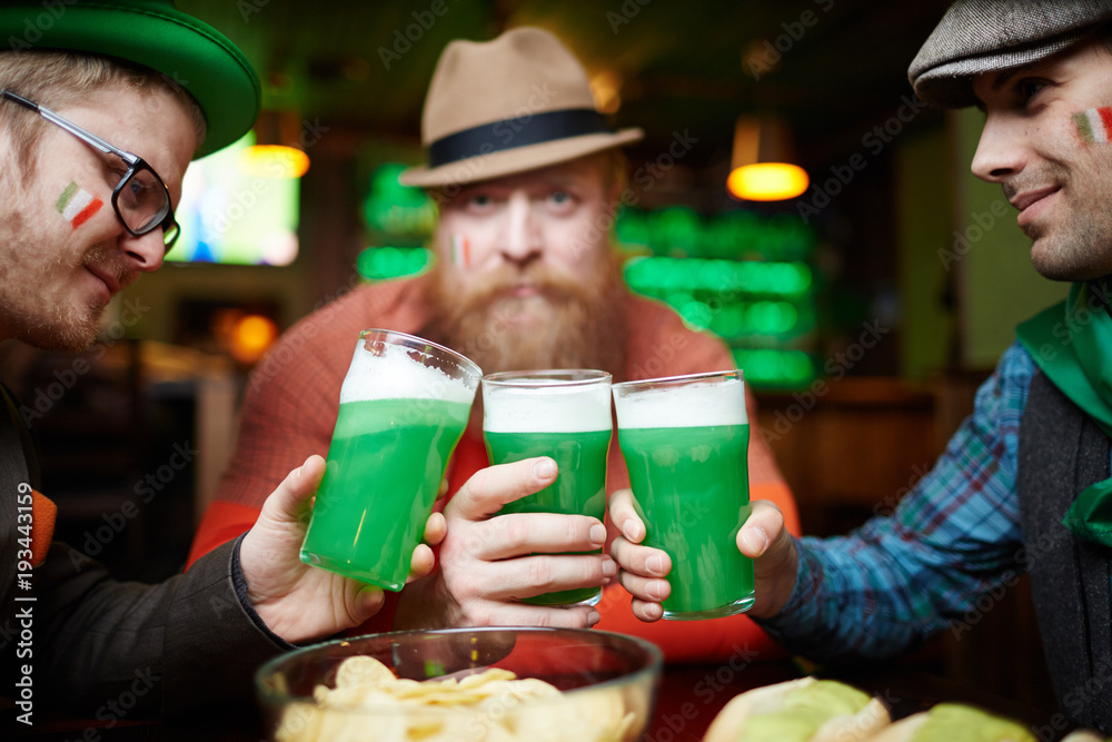 Friendly men toasting with glasses of beer over bowl with potato chips while spending time in bar