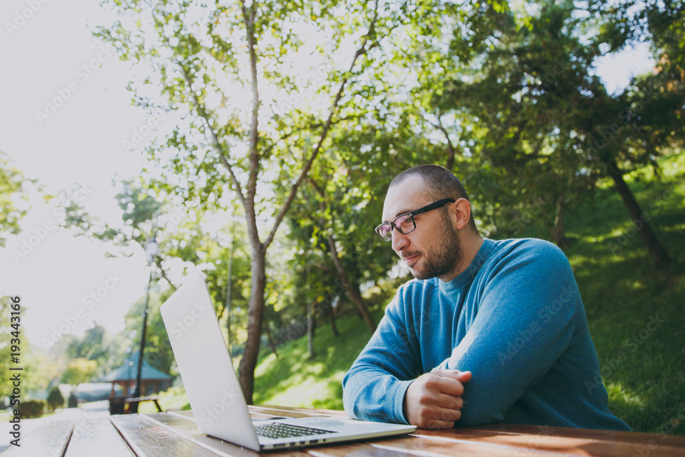 Young successful smart man businessman or student in casual blue shirt glasses sitting at table with mobile phone in city park using laptop working outdoors on green nature. Mobile Office concept.