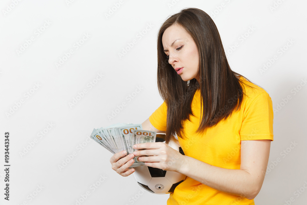 European young perplexed woman, football fan or player in yellow uniform counting bundle cash dollars soccer ball isolated on white background. Sport, play football game, excitement lifestyle concept. European young perplexed woman, football fan or player in yellow uniform counting bundle cash dollars soccer ball isolated on white background. Sport, play football game, excitement lifestyle concept.
