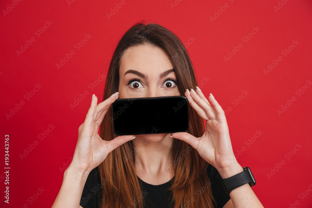 Amusing woman with long brown hair doing commercial and presenting new mobile device holding at face, isolated over red background Amusing woman with long brown hair doing commercial and presenting new mobile device holding at face, isolated over red background