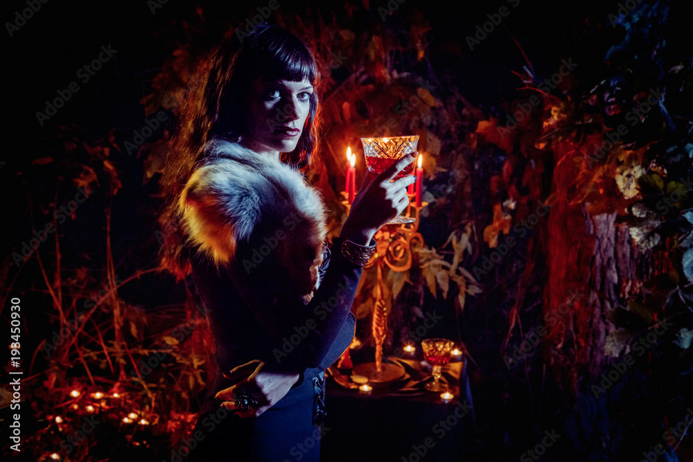 Beautiful witch with the glass of vine on the night forest background.