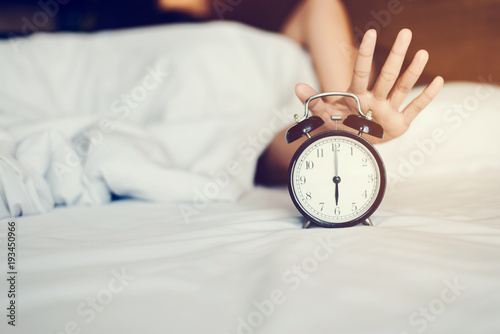 The woman's hand under the blanket reaches out for the alarm clock.