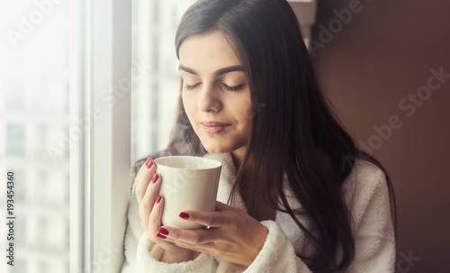 Beautiful girl wears white bathrobe smells cup of coffee sitting on the windowsill near window