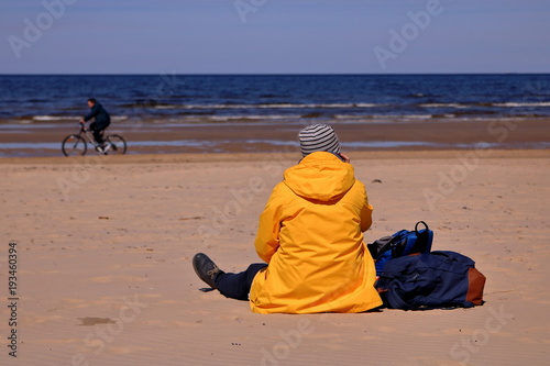 Samotna kobieta siedzi tyłem na piasku na plaży, w żółtej kurtce, obok niej leżą torby, w tle nieostry rowerzysta jedzie brzegiem morza, słonecznie photo