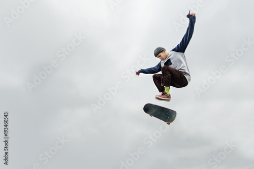 A skater teenager in a hat and a sweatshirt does a trick in the air with a flip of the board against a cloudy gray sky. The skater is isolated from other objects against the sky