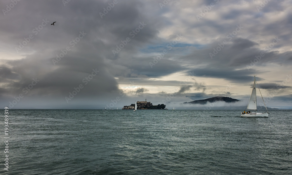 Scenic ocean landscape view with sailboat and small island covered in fog in background .Dark and misty storm clouds .