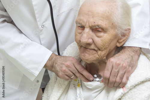 The internist doctor in a white dressing gown examines the lungs of a very old gray woman with a stethoscope. An internist doctor examines the lungs of a very old gray-haired woman sitting in a chair 