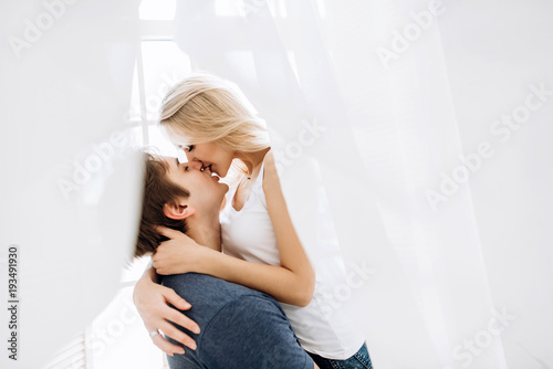 Amazing loving spouses kissing and has fun standing near the window in their bedroom, a husband holding his beloved beautiful wife in his arms