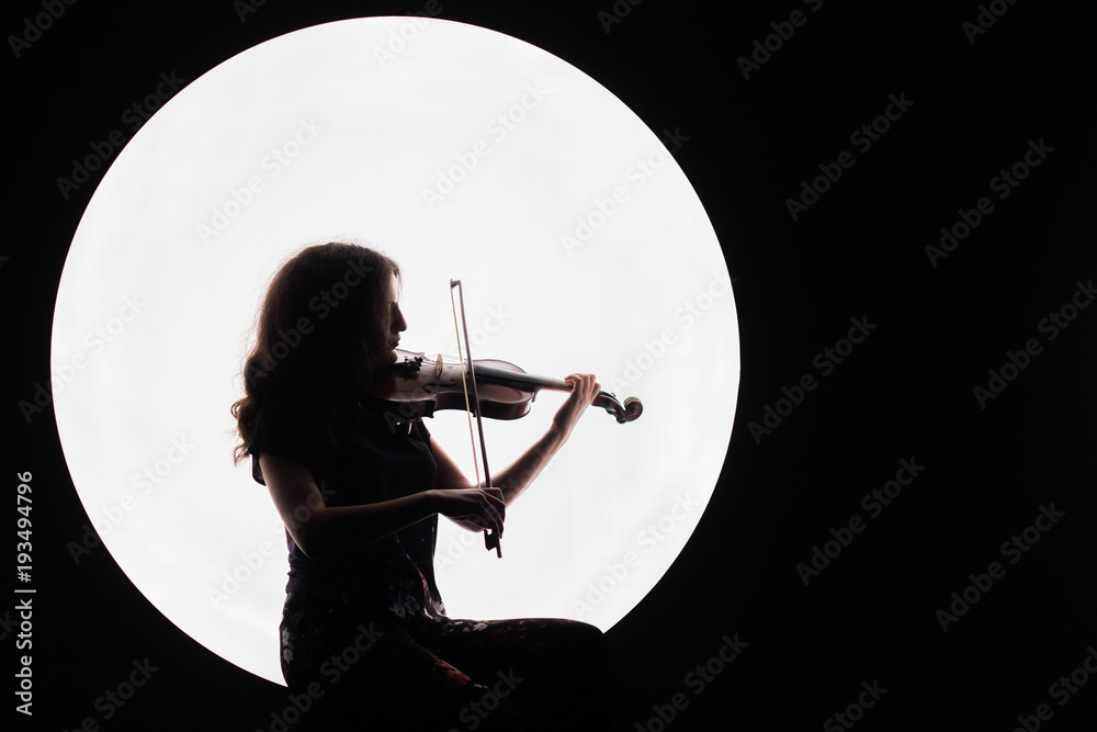 Fototapeta Silhouette of a beautiful brunette girl playing the violin. Concept for music news. Copy space. White circle as the moon on a black background.