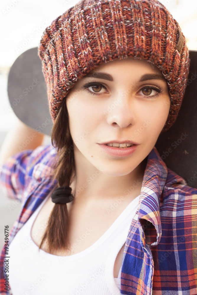 Brunette teenage girl in hipster outfit (jeans shorts, keds, plaid shirt, hat) with a skateboard at the park outdoors Brunette teenage girl in hipster outfit (jeans shorts, keds, plaid shirt, hat) with a skateboard at the park outdoors