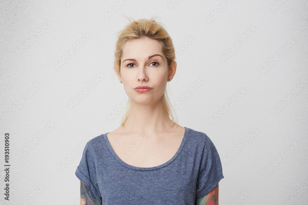 What's going on? Confused and surprised girl, whith tattoos over her arms and pierced nose do not understand and raise her eyebrow. Serious and mad. Isolated over white wall