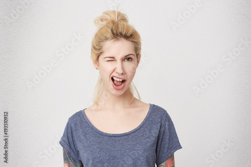 Portrait og young stylish blonde girl laughs and wink, wears blue casual t-shirt. Emotional expression. Positive. Isolated over white wall © timtimphoto
