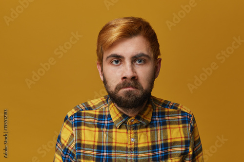 Young bearded male feels angry and mad about some problems and people. Wearing shirt, blue eyes and dyed blond hair. Isolated over yellow background.