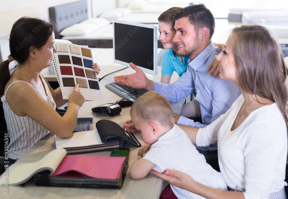 Fototapeta premium Smiling seller female is showing to young family the patterns of mattress fabrics