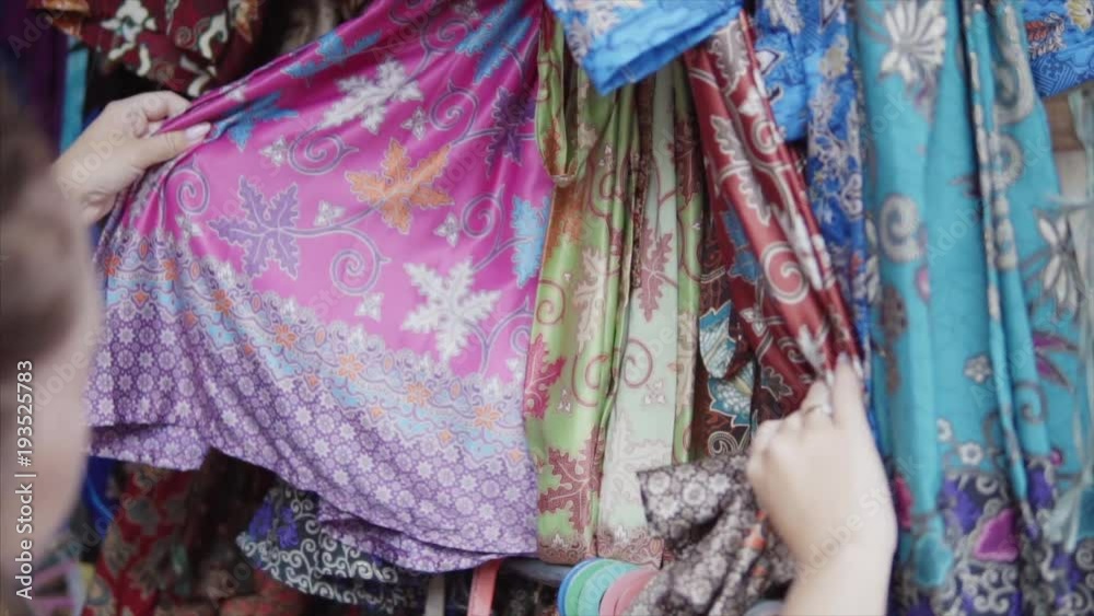 Stand of the colorful embroidered with threads and stamp print clothes in different colours at the traditional etnic asian market. Womans hand trying the quality of the textile material Stand of the colorful embroidered with threads and stamp print clothes in different colours at the traditional etnic asian market. Womans hand trying the quality of the textile material