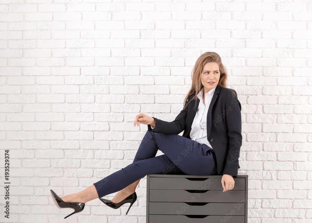 Φωτογραφία Beautiful fashionable woman in elegant suit sitting on chest of drawers against