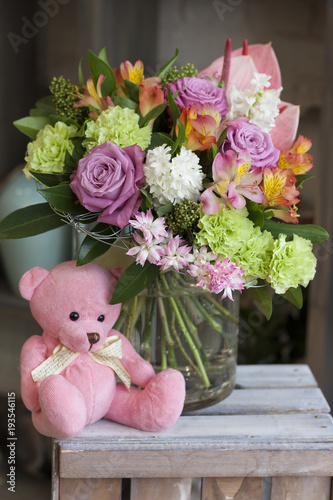 Розовый игрушечный мишка сидит на деревянном ящике рядом с букетом цветов