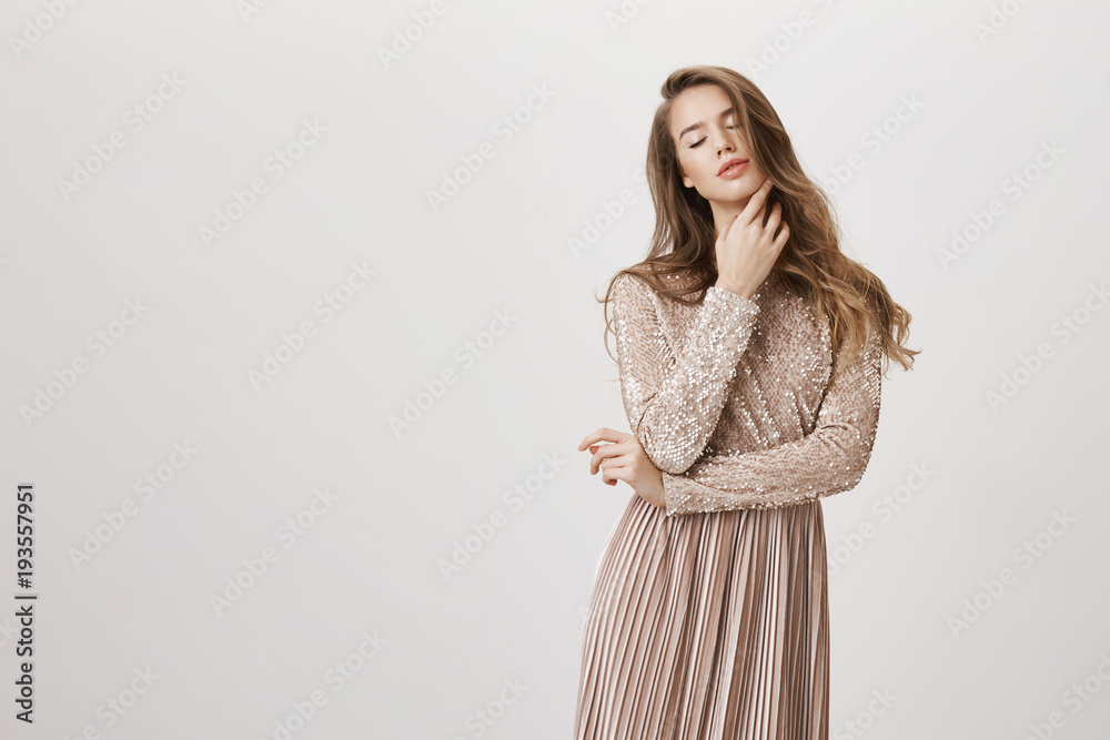 Sensual feminine caucasian female model standing in beige trendy evening dress, softly touching skin while standing with closed eyes over gray background. Tender girlfriend dreams about love Sensual feminine caucasian female model standing in beige trendy evening dress, softly touching skin while standing with closed eyes over gray background. Tender girlfriend dreams about love