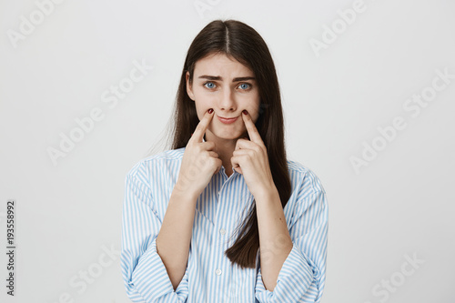 Indoor portrait of unhappy and stressed woman with frowned eyebrows stretching mouth with index fingers to make gloomy smile, standing over gray background. Office clerk tired of constantly smiling