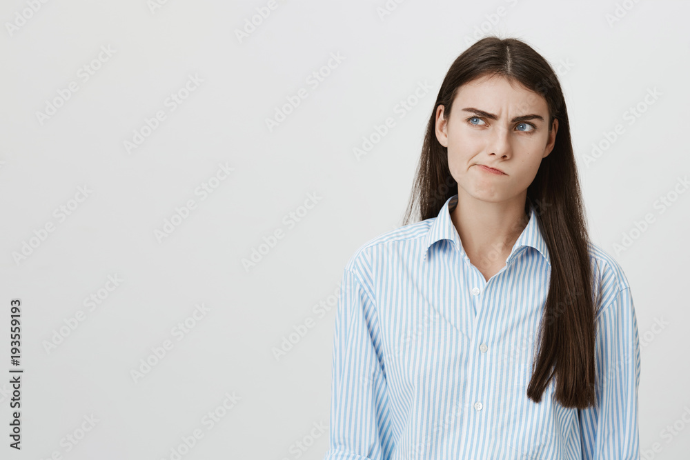 Portrait of interesting european female smirking while thinking about something and looking aside with frowned eyebrows, standing over gray background. Student making up excuse why she was absent Portrait of interesting european female smirking while thinking about something and looking aside with frowned eyebrows, standing over gray background. Student making up excuse why she was absent