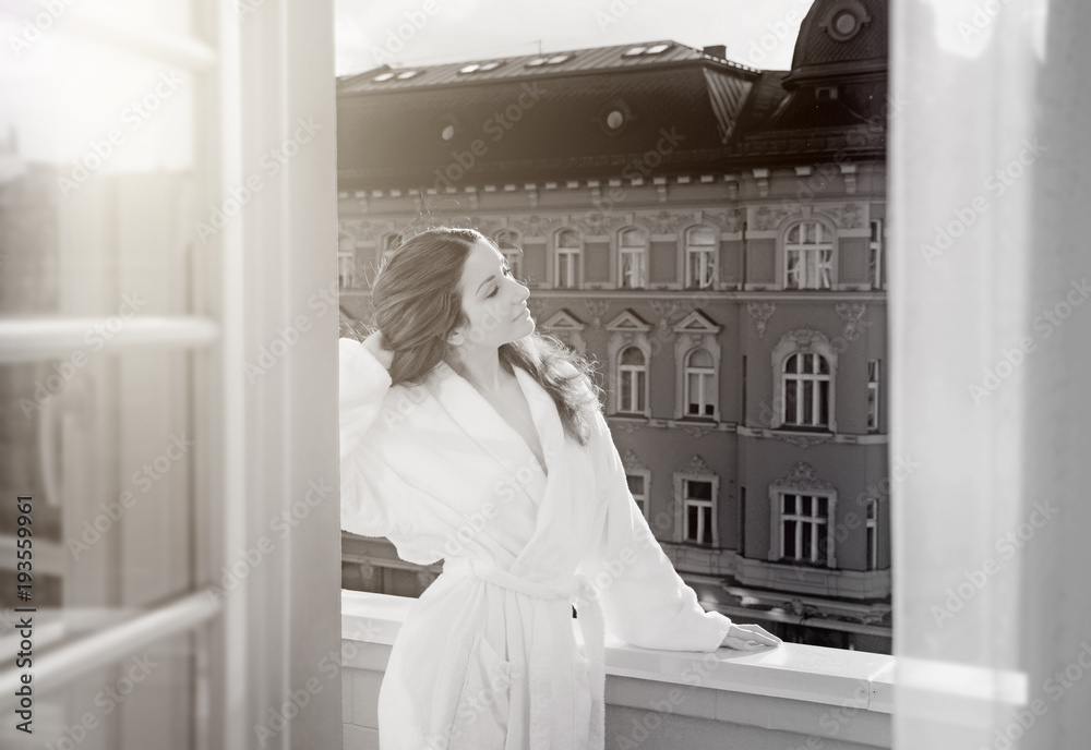 Fototapeta premium Beautiful woman at morning wearing robe standing on the balcony overlooking old European streets. Concept of beautiful life, relax or vacation 