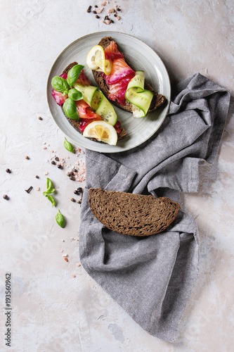 Sliced beetroot marinated salmon sandwiches with rye bread, cucumber, basil and lemon served on ceramic plate with textile napkin over grey texture background. Top view, space photo