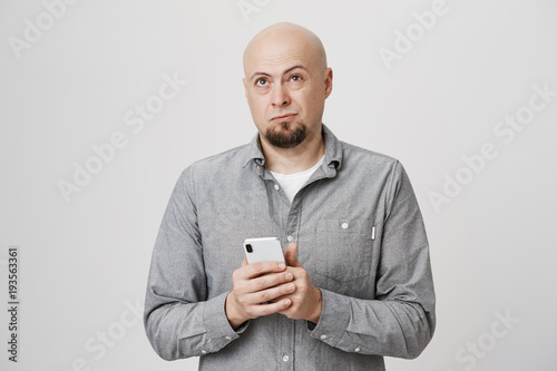Φωτογραφία Portrait of handsome bald european guy holding smartphone while frowning and looking up, trying to recall phone number, standing over gray background