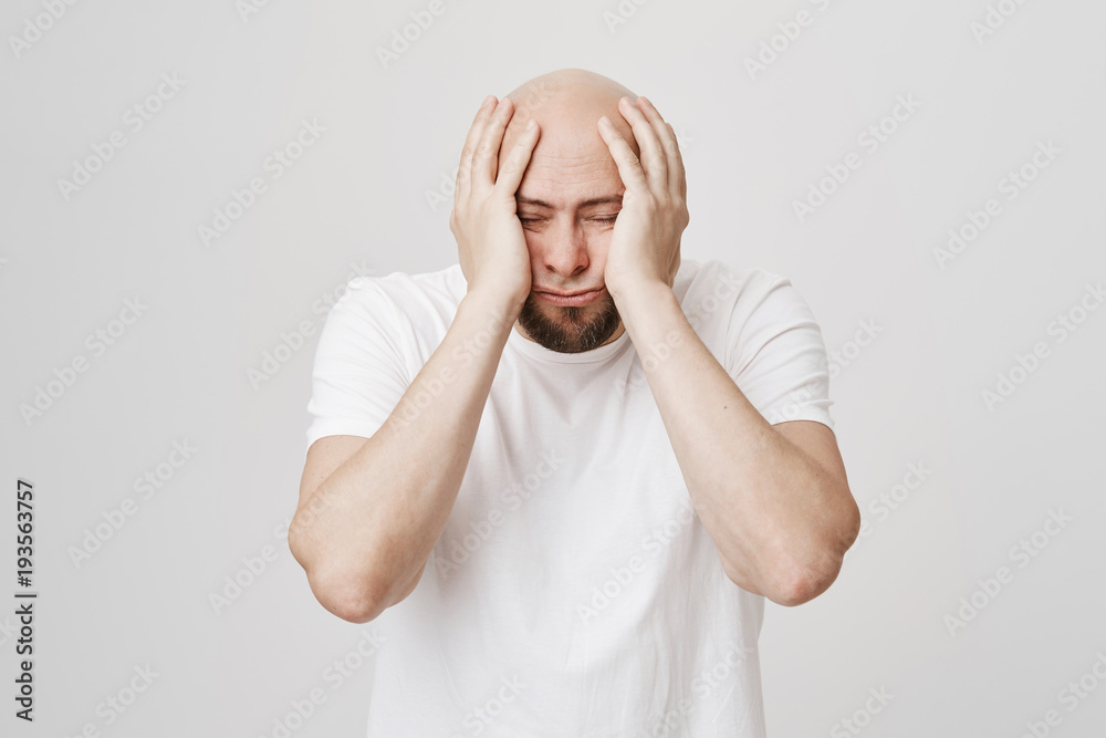 Portrait of tired or stressed bald caucasian male holding hands on head, squeezing face and standing with closed eyes over gray background. Guy have not slept at all and has lots of work Portrait of tired or stressed bald caucasian male holding hands on head, squeezing face and standing with closed eyes over gray background. Guy have not slept at all and has lots of work