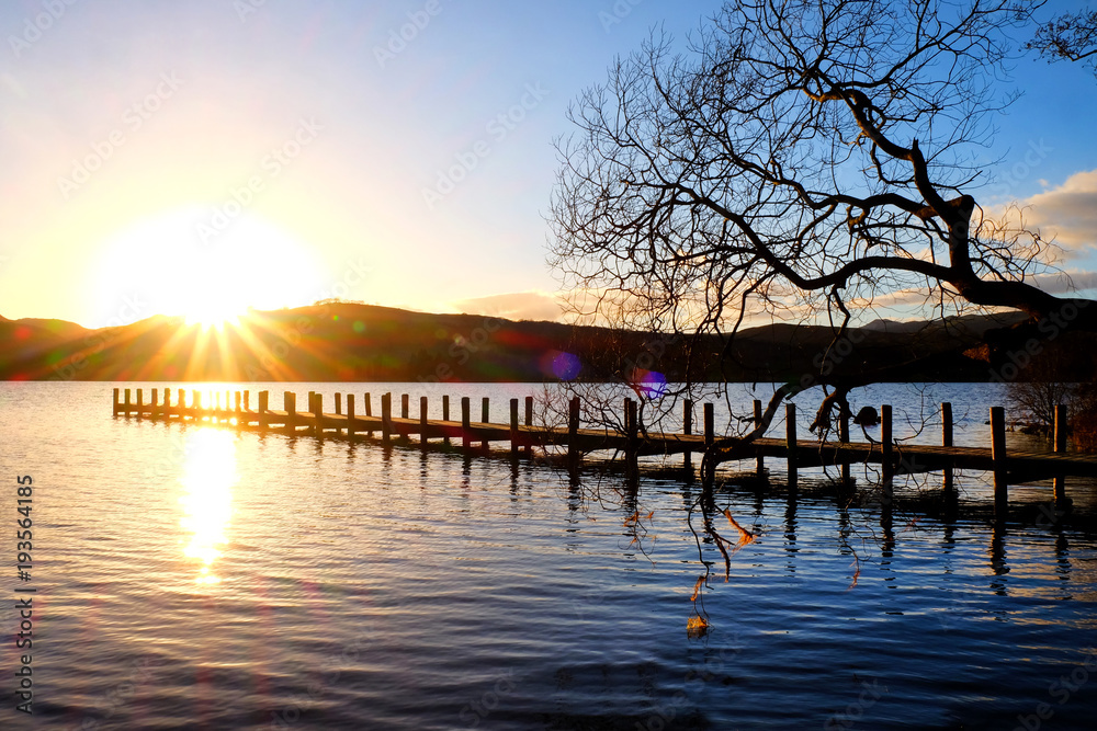 Fototapeta premium very long wooden jetty, jutting out into a calm blue wooden lake with mountains sunsetting in background