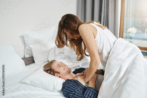 Indoor shot of happy laughing boyfriend lying on bed while his girlfriend sits on him and touches his chest. Couple messing around in bedroom. Wife wakes up her husband.