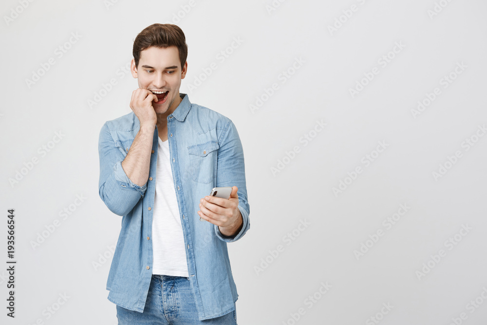 Emotive young cute student with surprised and amazed expression looking at smartphone biting fingers and smiling, isolated over white background. Man excited to video chat with friend who lives abroad Emotive young cute student with surprised and amazed expression looking at smartphone biting fingers and smiling, isolated over white background. Man excited to video chat with friend who lives abroad