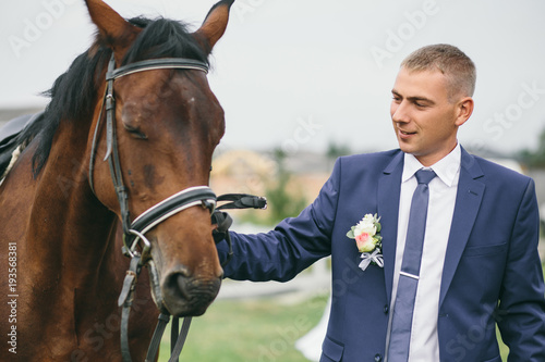 Handsome groom in blue suit poses with brown horse outside