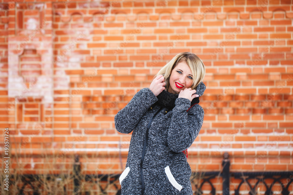 A beautiful hipster girl in a gray coat smiles and looks at the camera on a red background outdoors. Portrait of a modern girl with red lips against a brick wall background.