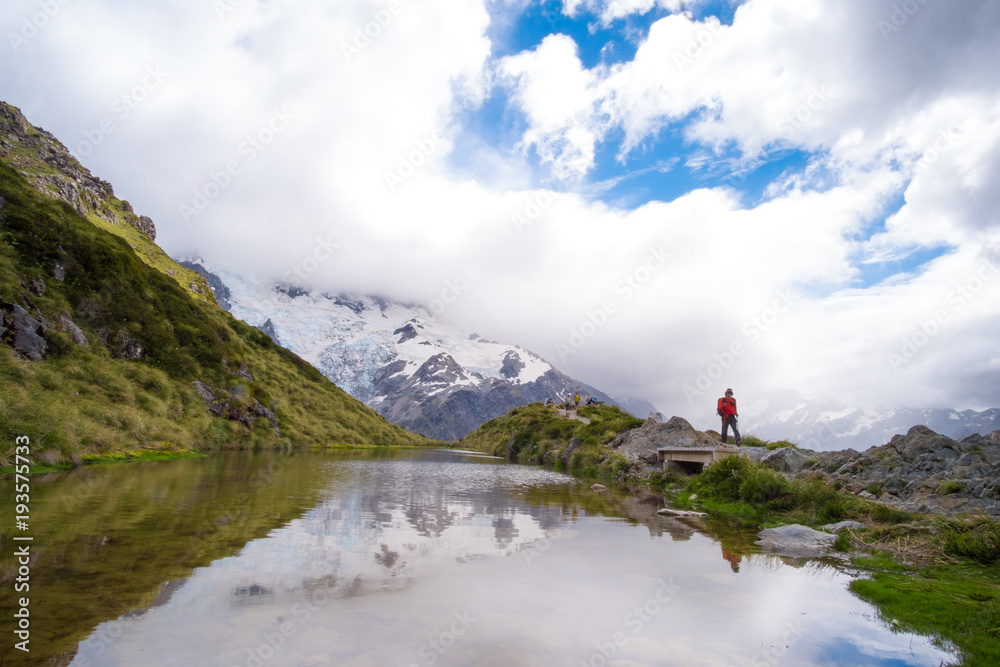 The reflection of the rocky mountain with snow and blue sky with cloud and traveller on the small lake on the high mountain in Mt Cook National Park (Muller hut track)