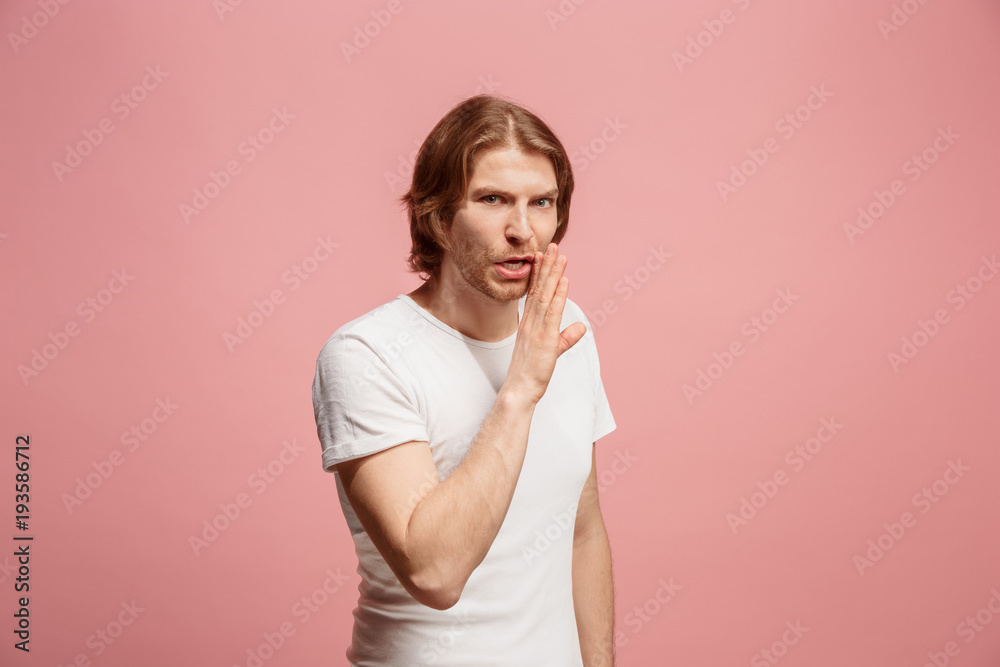 The young man whispering a secret behind her hand over pink background The young man whispering a secret behind her hand over pink background