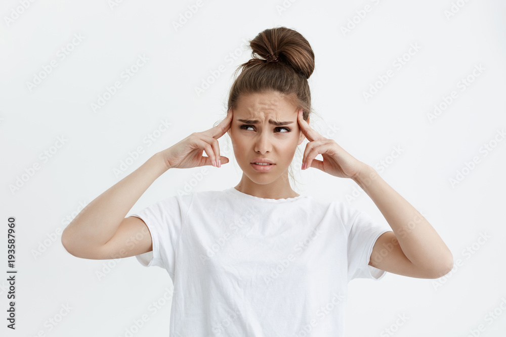Indoor shot of perplexed and disappointed cute woman holding fingers on temples, looking aside with worried expression, feeling headache or being unable to focus, standing over white background. Indoor shot of perplexed and disappointed cute woman holding fingers on temples, looking aside with worried expression, feeling headache or being unable to focus, standing over white background.