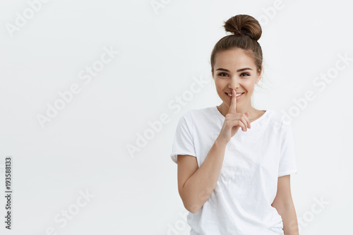 Cheerful fashionable european female smiling while showing shh gesture with index finger on lips, looking with positive expression and standing over white background. Secret and body language concept