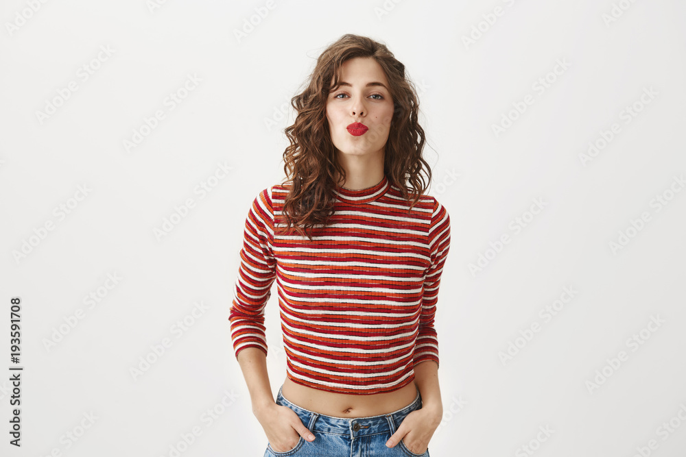 Come and save me prince charming. Studio shot of gorgeous caucasian model with curly hairstyle and red lipstick folding lips while waiting for kiss, standing with hands in pockets and sensual look