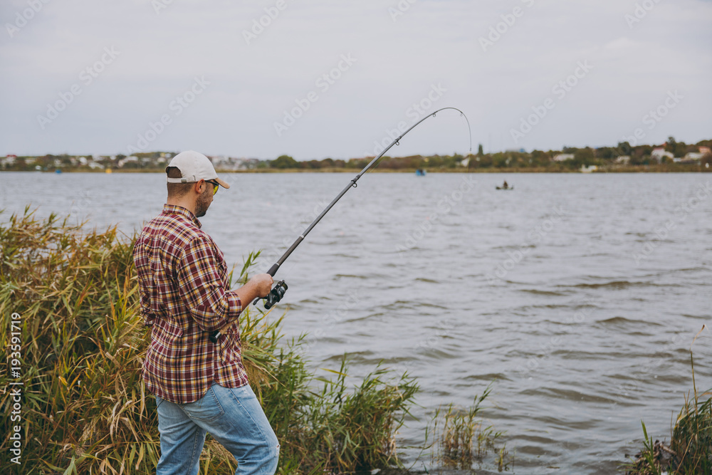 Side view Young unshaven man with a fishing rod in checkered shirt, cap and sunglasses casts fishing pole on a lake from shore near shrubs and reeds. Lifestyle, recreation, fisherman leisure concept Side view Young unshaven man with a fishing rod in checkered shirt, cap and sunglasses casts fishing pole on a lake from shore near shrubs and reeds. Lifestyle, recreation, fisherman leisure concept