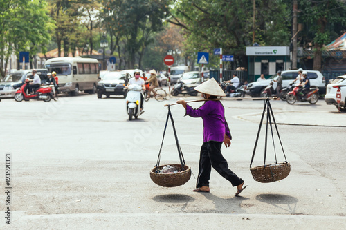 Woman wearing purple cloth and conical hat. Carry baskets with stuff inside with a carrying pole on the street in Hanoi, Vietnam.

