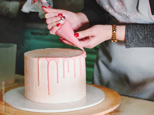 Фотография Unrecognisable woman decorating mousse glaze cake with rose, macarons, hands detail, focus on the cake