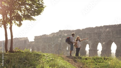 Young lovely couple backpackers tourists reading map pointing directions roman aqueduct arches in parco degli acquedotti park ruins in rome on romantic misty sunrise with guitar and sleeping bag slow photo