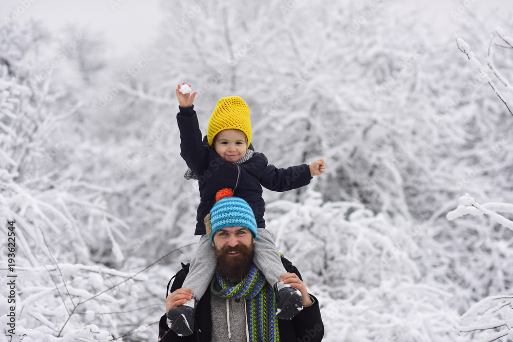 Bearded young father and beautiful little son in bright knitted hats on walk in winter forest. Boy sits in man shoulders and holds a snowball in raised up hand. Funny people dressed in bright clothes Bearded young father and beautiful little son in bright knitted hats on walk in winter forest. Boy sits in man shoulders and holds a snowball in raised up hand. Funny people dressed in bright clothes
