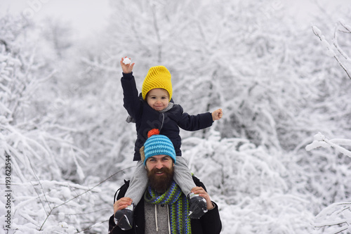 Bearded young father and beautiful little son in bright knitted hats on walk in winter forest. Boy sits in man shoulders and holds a snowball in raised up hand. Funny people dressed in bright clothes