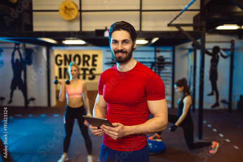 Portrait of handsome male personal trainer with tablet. Blurred girl in the background doing workout.