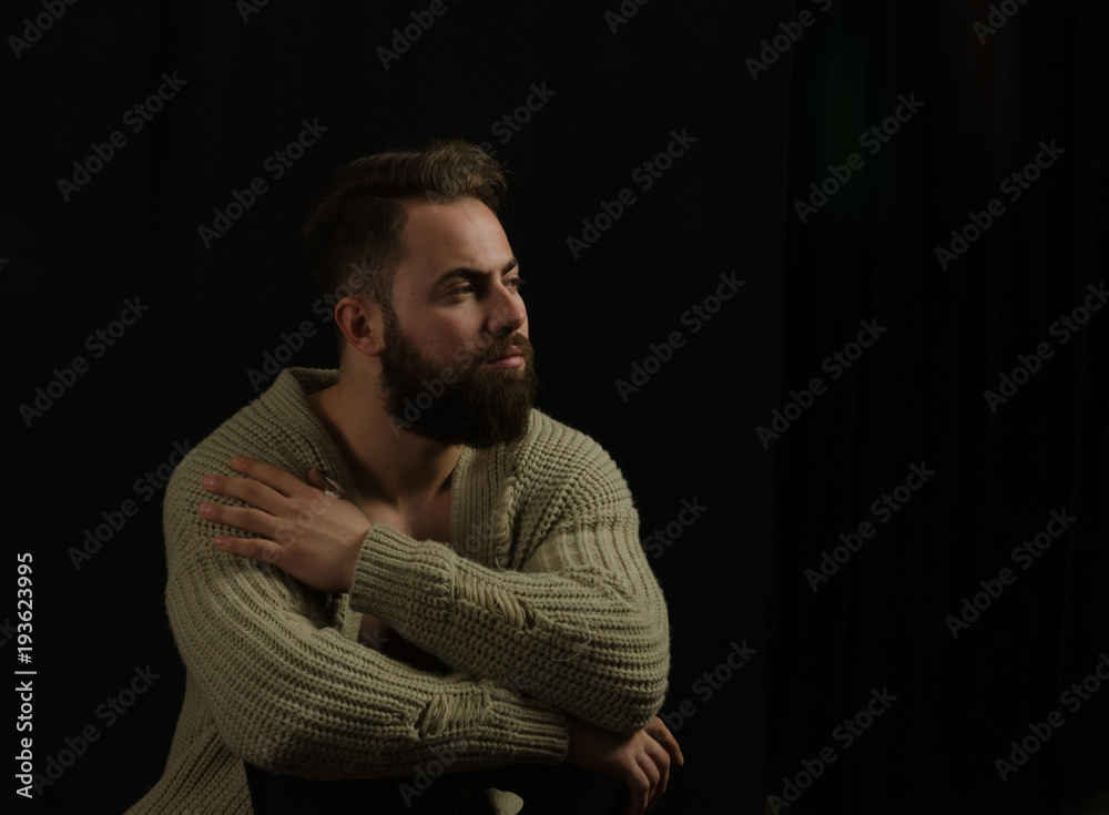 The young model posing in the studio is dressed in a cardigan and is in his hand pockets and bearded ... sitting on a chair, looking ahead. The young model posing in the studio is dressed in a cardigan and is in his hand pockets and bearded ... sitting on a chair, looking ahead.