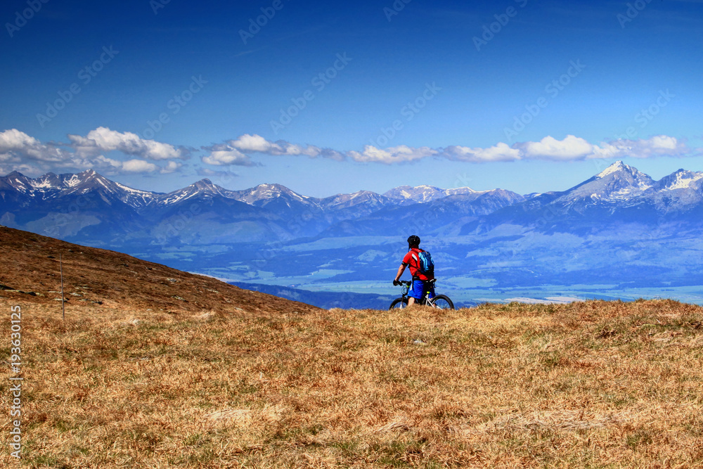 Biker rides a mountain bike in popular hiking area sunlit Low Tatras meadow with snowy jagged Zapadne and Vysoke Tatry ranges highest of Slovakia in background