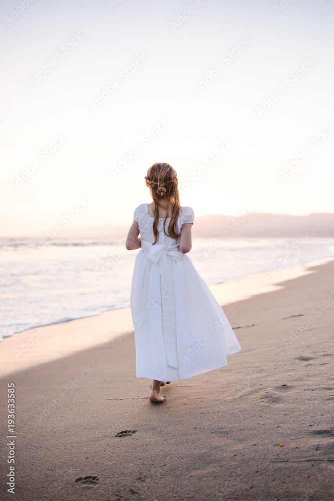 Young girl in white dress walking away from the camera in the sand along the waterline of the beach Young girl in white dress walking away from the camera in the sand along the waterline of the beach