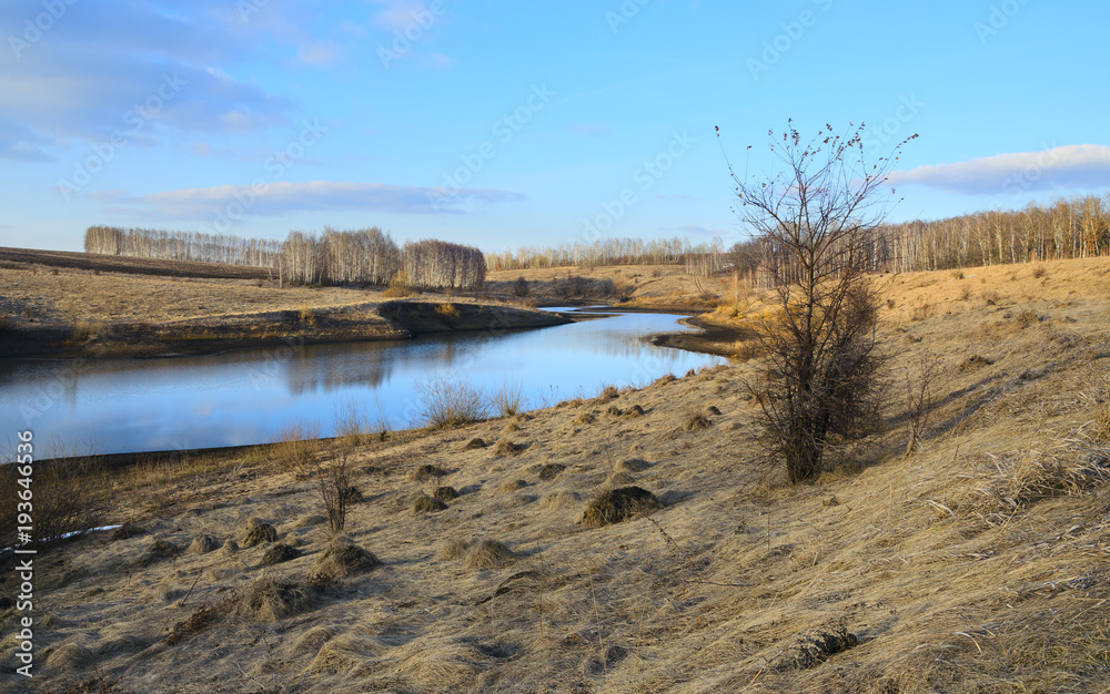 Springtime.Sunny spring colorful landscape with river and bare birches growing on the riverbank.Warm sunlight at sunset.Clouds in blue sky.Beautiful view. 