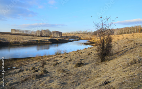 Fototapeta Naklejka Na Ścianę i Meble -  Springtime.Sunny spring colorful landscape with river and bare birches growing on the riverbank.Warm sunlight at sunset.Clouds in blue sky.Beautiful view. 